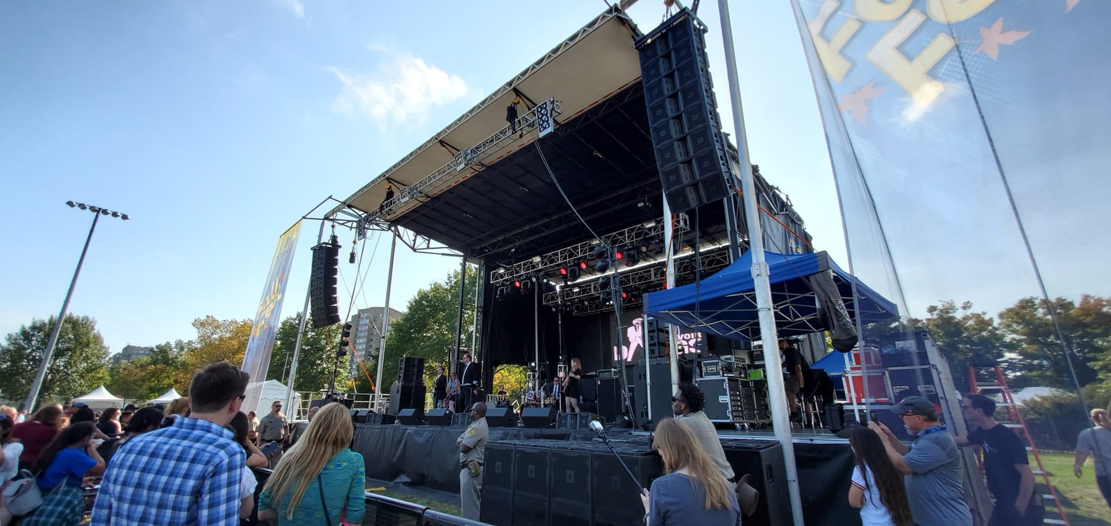 An outdoor festival stage with flown line-array speakers and a band playing, seen from the crowd.