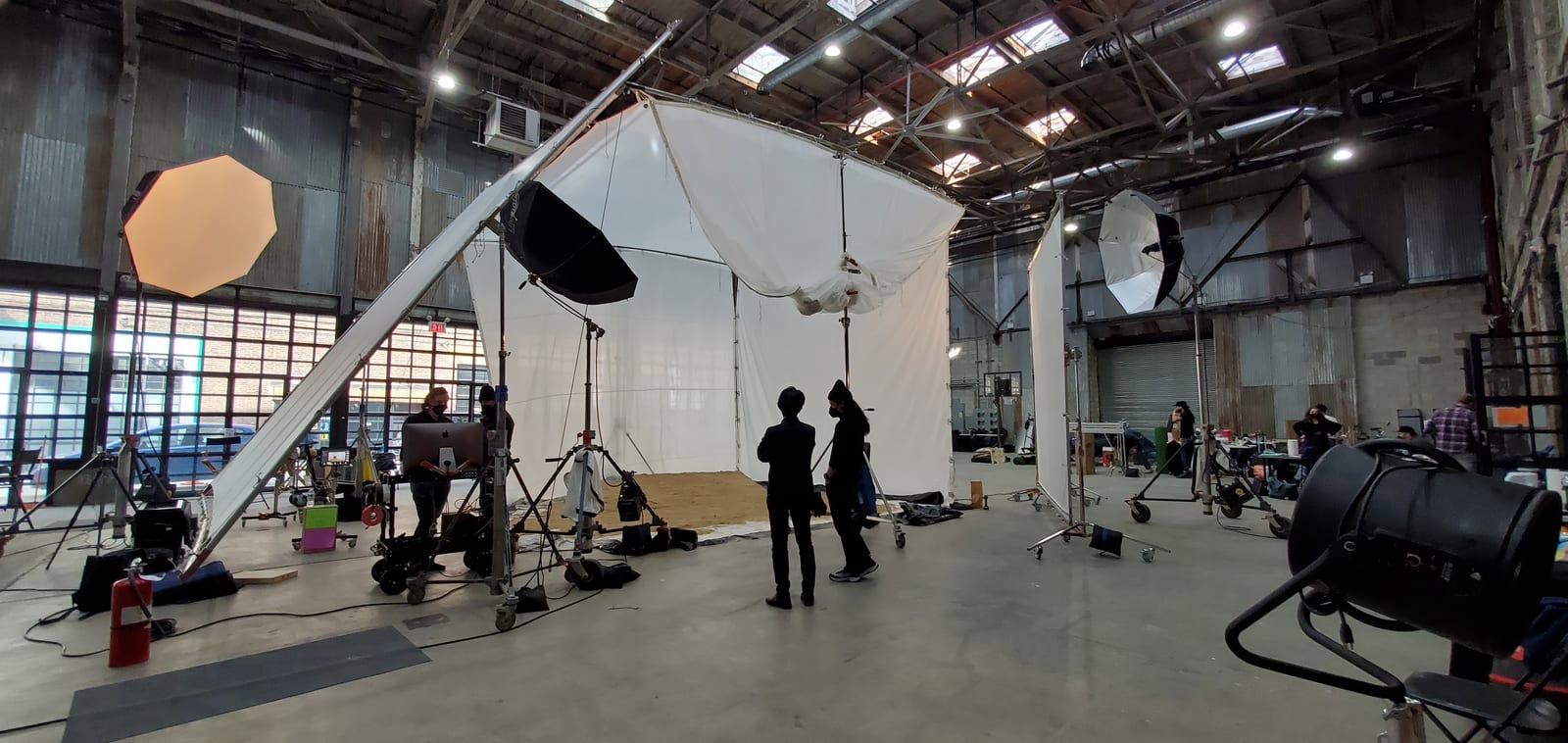 Crew rigging a white cyclorama, softboxes and grip stands inside a warehouse soundstage.