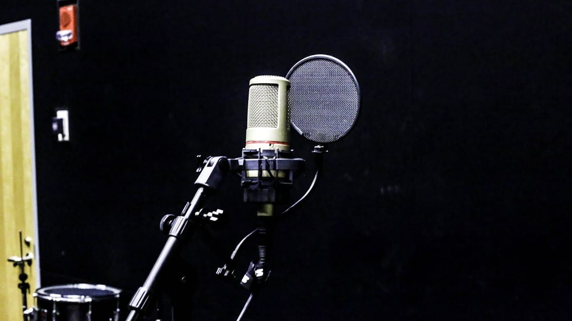 A large-diaphragm condenser microphone and pop filter on a boom stand in a darkened studio, with a snare drum just in frame.